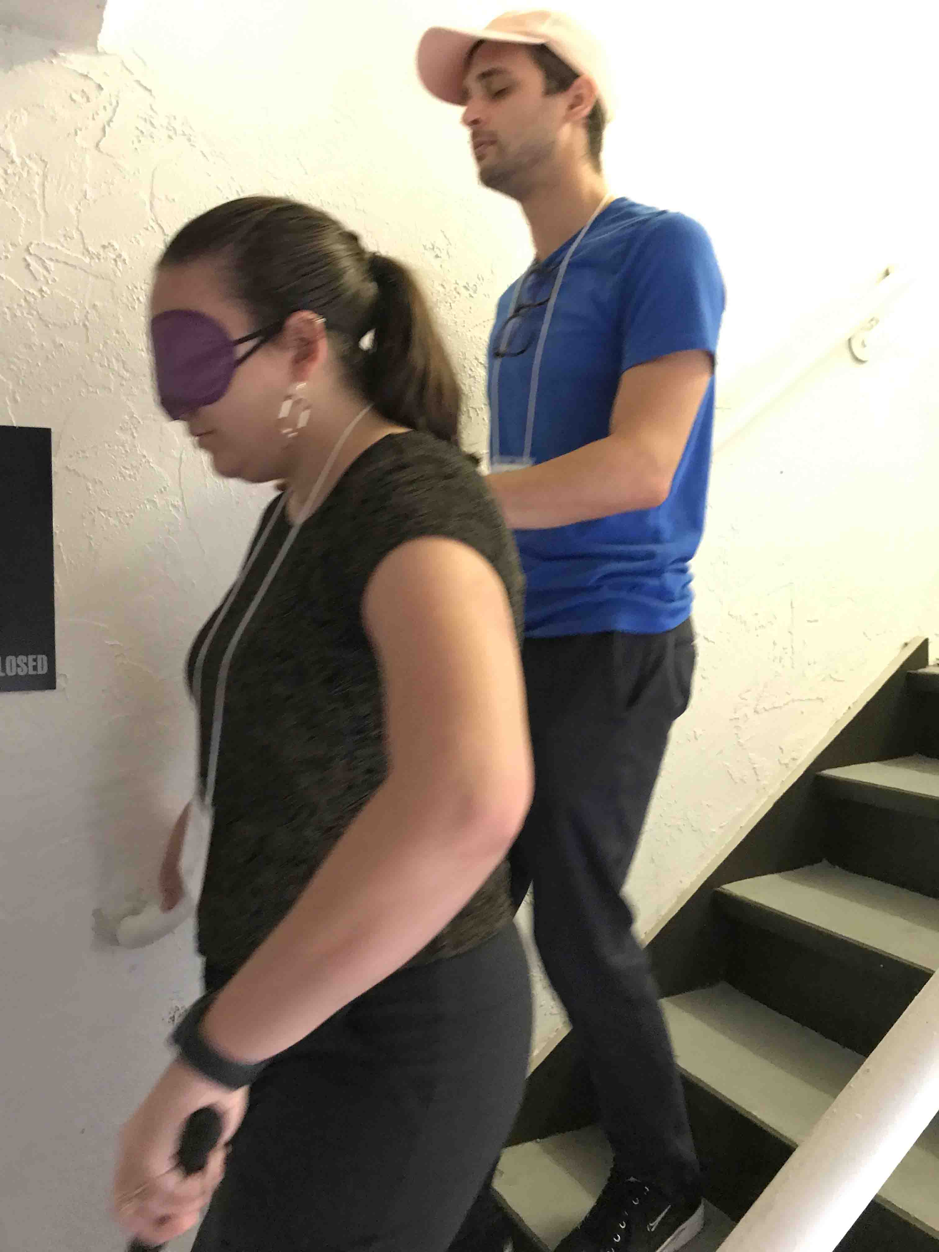 2 pictures show a woman wearing a blindfold and holdng a cane in her left hand and the railing in her right hand as she descends a stairway.  She is guiding a man on her right, he has his eyes closed and is holding the guide's right arm with his left hand, and the railing with his right hand as they descend.
